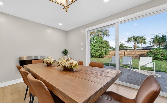 Modern dining room with wood table, tan leather chairs, brass pendant light, and sliding glass doors opening to backyard with palm trees