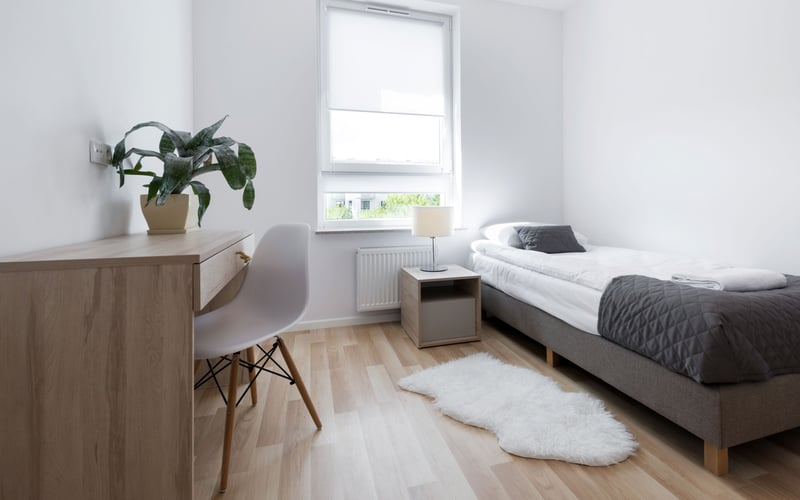 Bright bedroom with wooden desk, white chair, potted plant, bed with gray bedding, and window with radiator