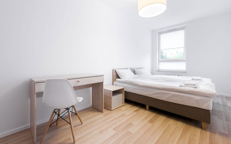 Bright bedroom with double bed, desk, white chair, wooden floor, and window with blinds