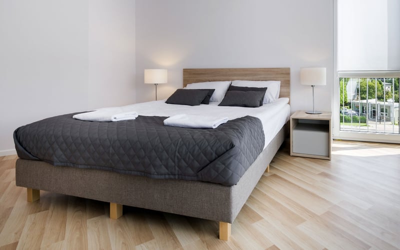 Bright bedroom with queen bed, black quilted comforter, wooden headboard, nightstands with lamps, and glass doors opening to garden.