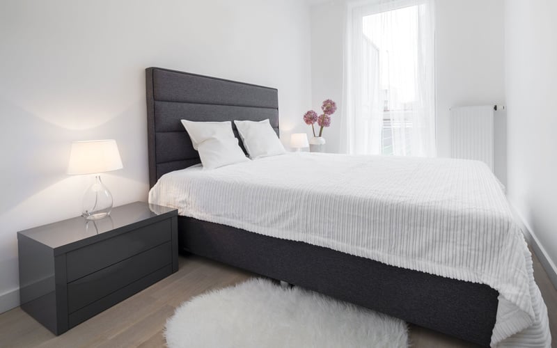 Bright bedroom with dark gray upholstered bed, white bedding, black nightstand with lamp, and pink flowers by window