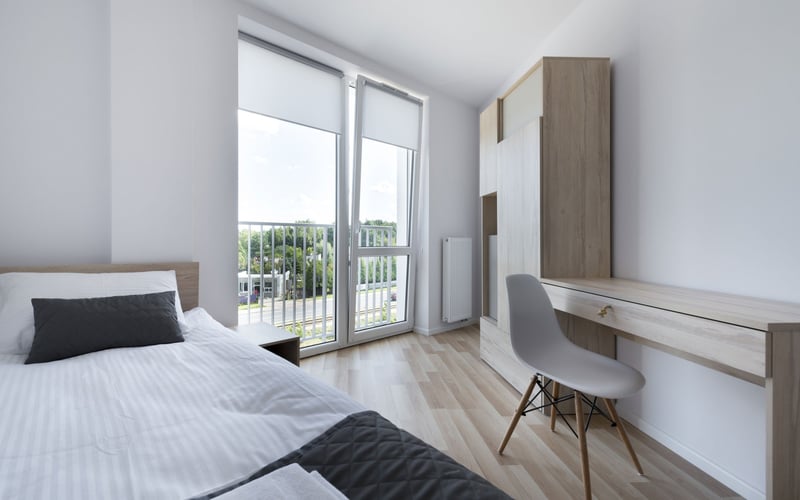 Modern bedroom with bed, desk, wardrobe, and glass doors opening to balcony with tree views